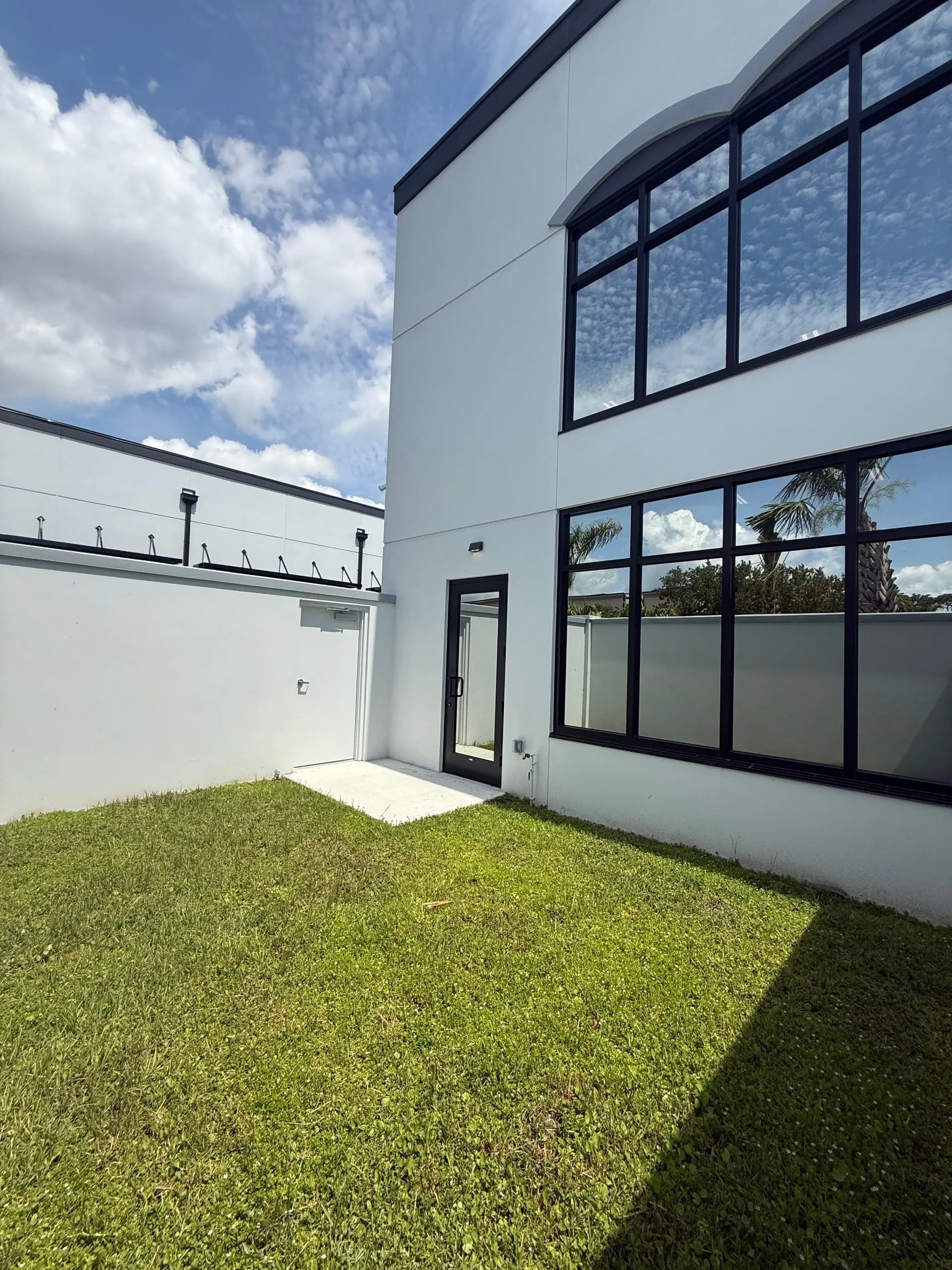 Modern white exterior with black-framed windows and glass door, private landscaped courtyard with grass and palm tree visible beyond