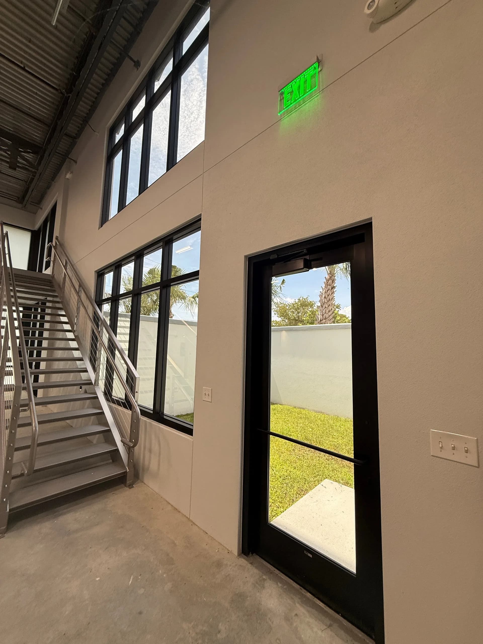 Mezzanine interior with metal staircase, beige walls, black-framed windows and glass door opening to landscaped grounds with palm trees, green EXIT sign visible
