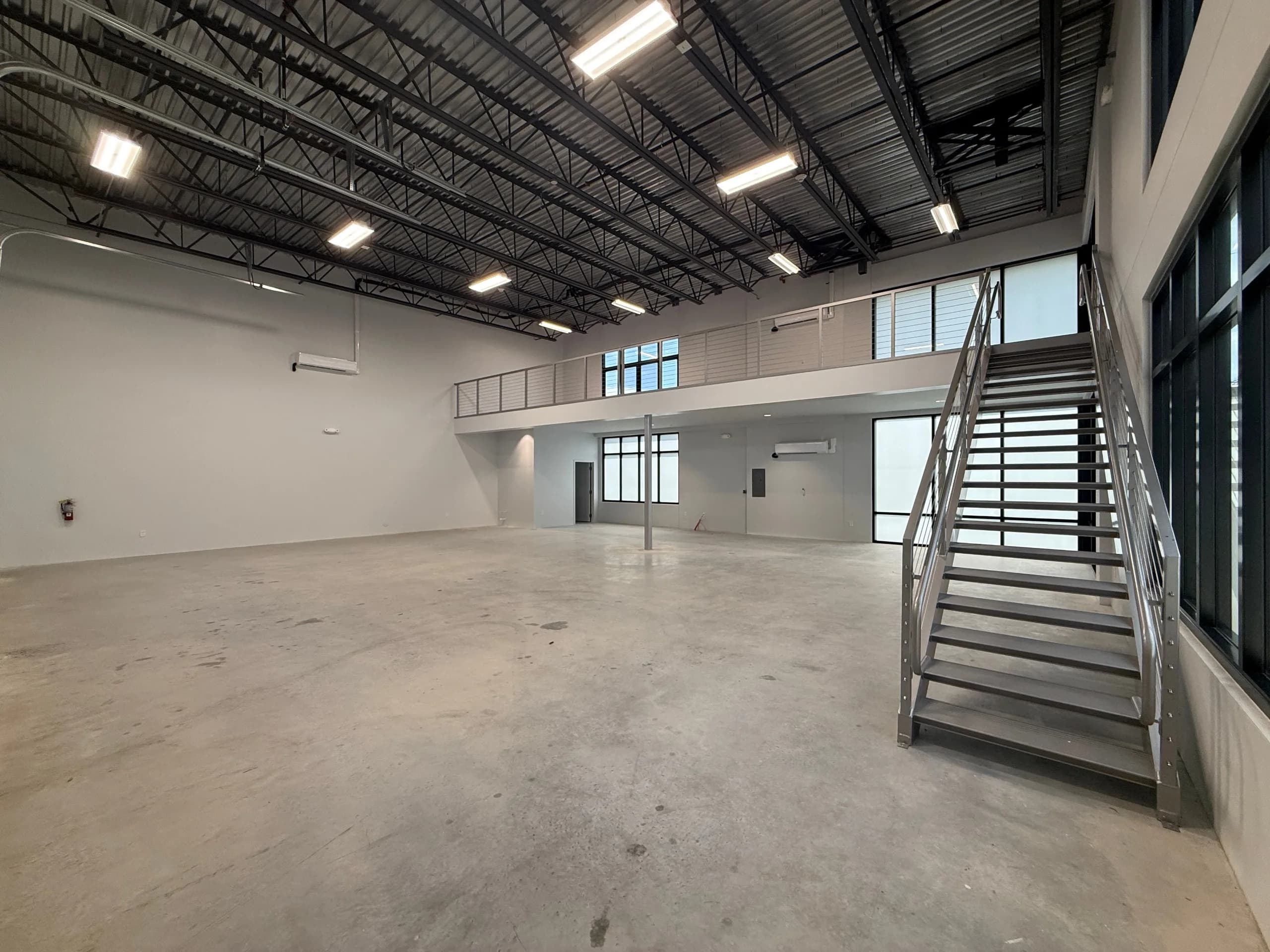 Large warehouse-style unit with concrete floor, white walls, metal staircase leading to mezzanine level with railing, expansive industrial ceiling with exposed steel trusses and LED lighting