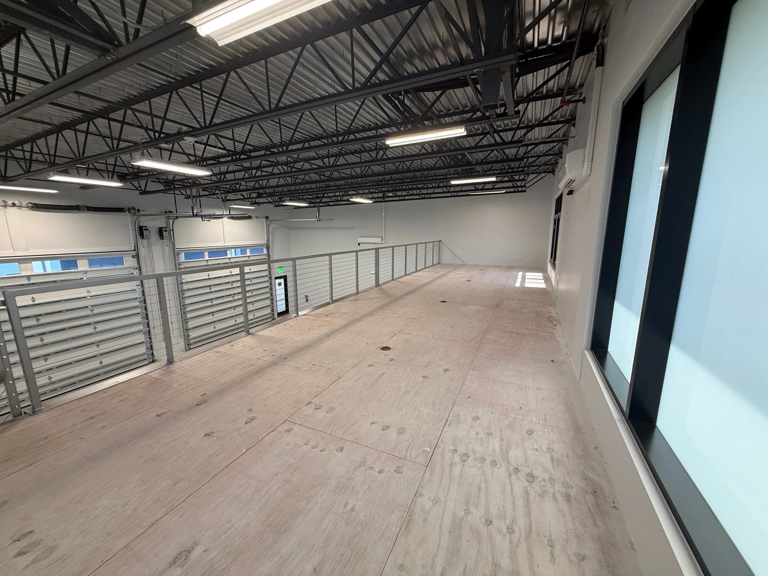 Spacious mezzanine level with wood-look flooring, white walls, black steel roof trusses, metal railing, and fluorescent lighting