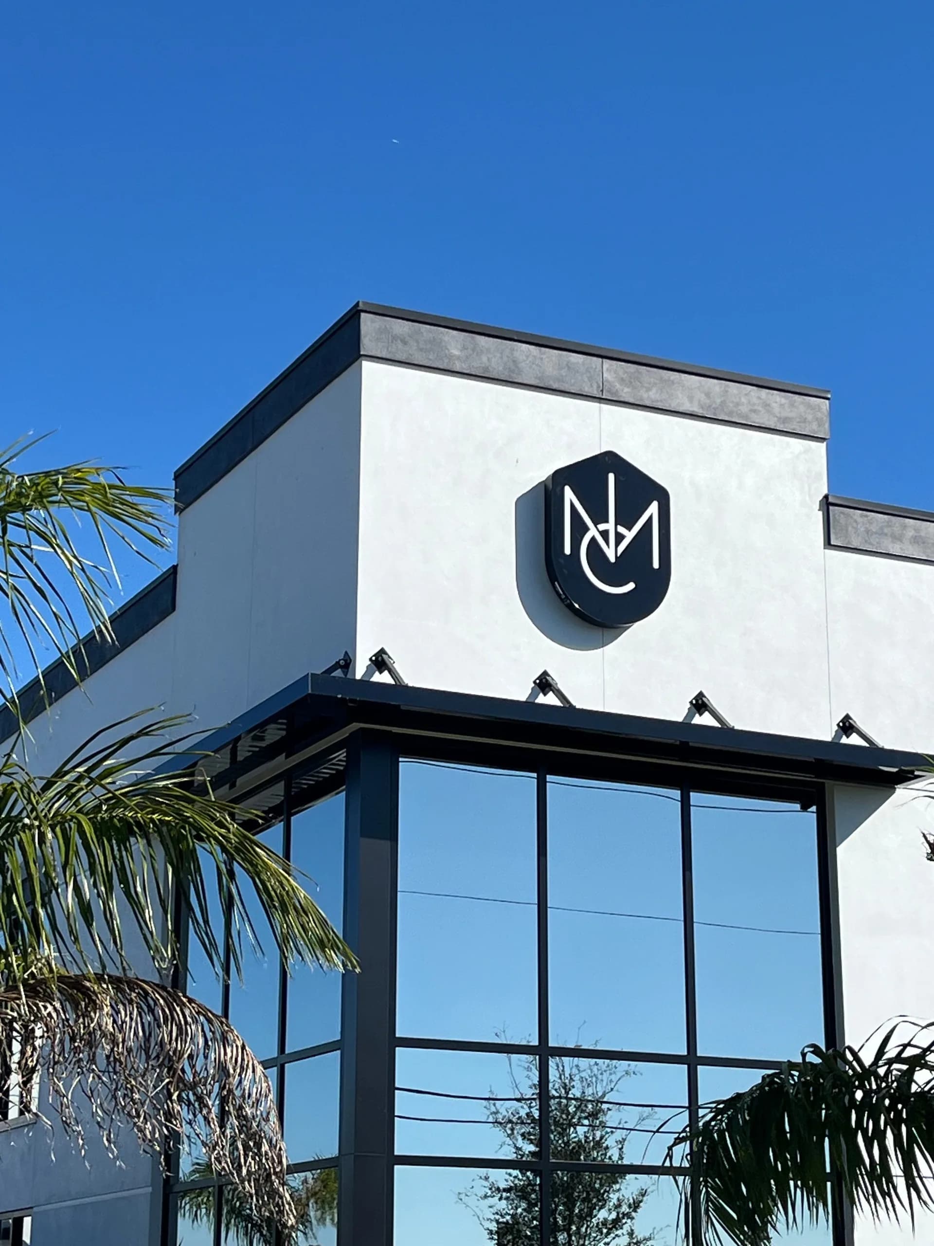 Modern white and black building exterior with logo signage, large glass windows, and decorative roof elements, flanked by palm fronds