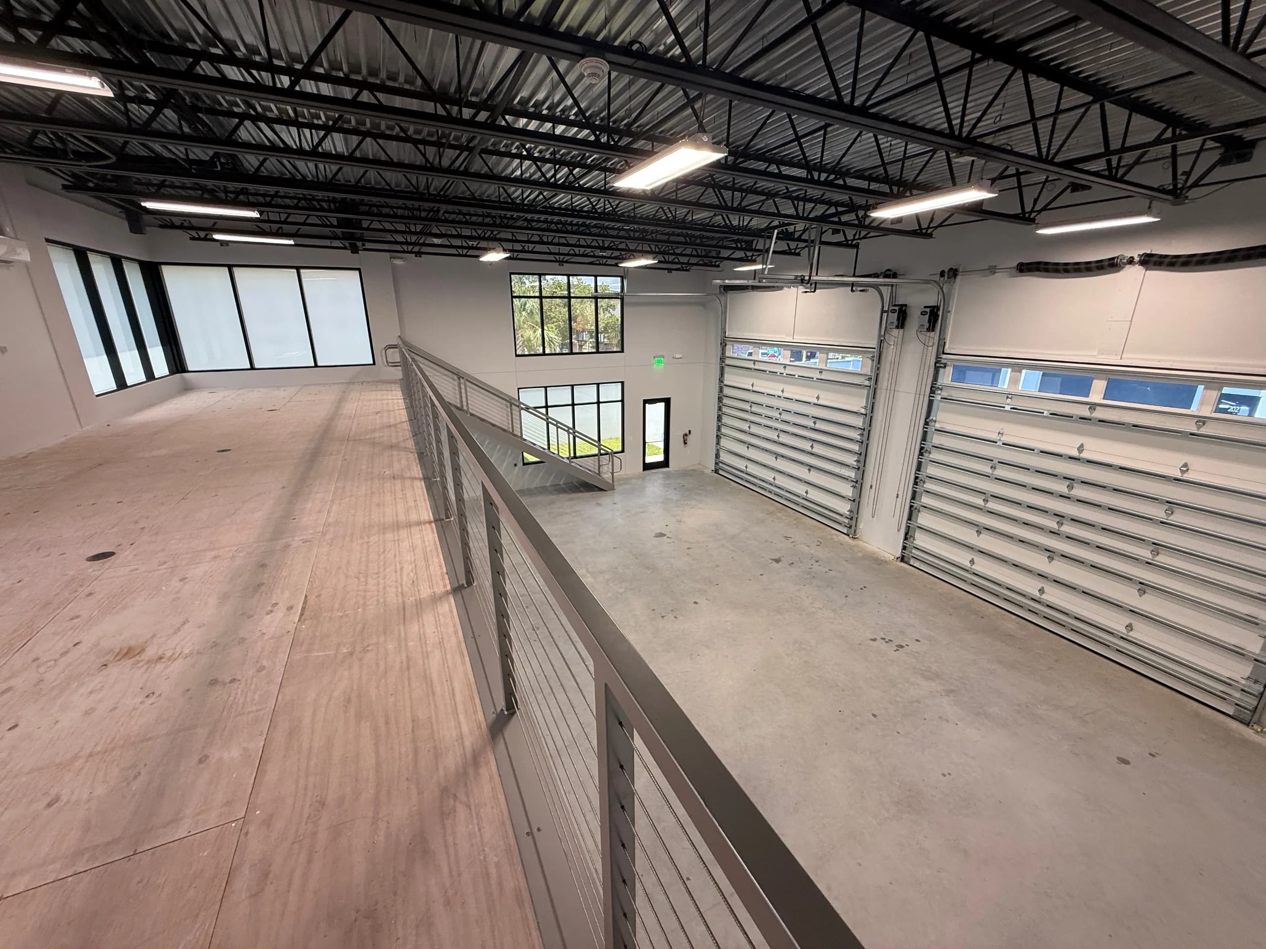Mezzanine level with plywood flooring and metal railings, metal-framed windows overlooking lower drive-in storage bays, exposed structural steel ceiling with LED lighting